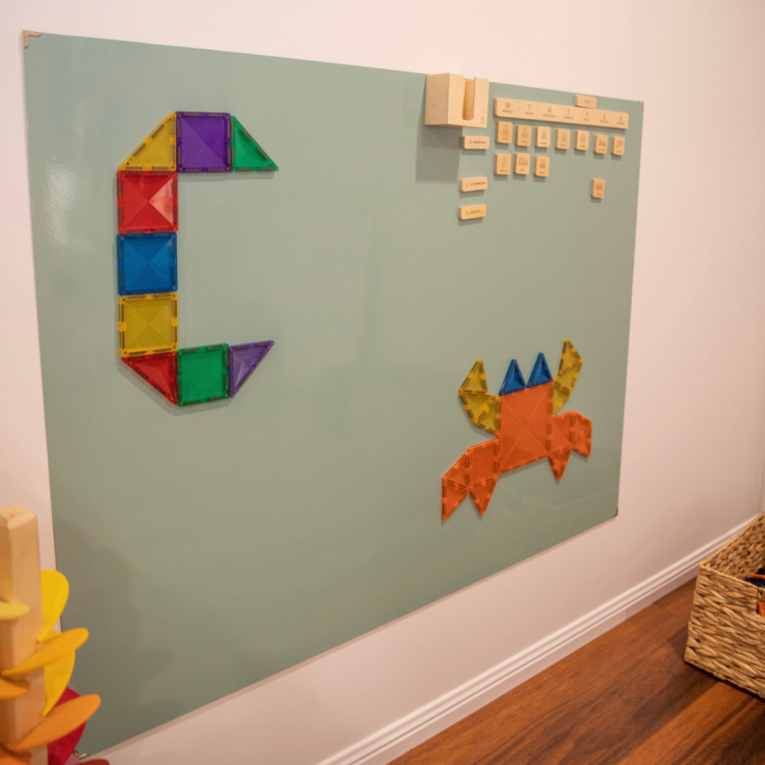 A pretty green "whiteboard" being used to play and display. There is the letter 'C' and a crab picture made of Connetix Magnetic Tiles. A beautiful feature wall of a playroom.